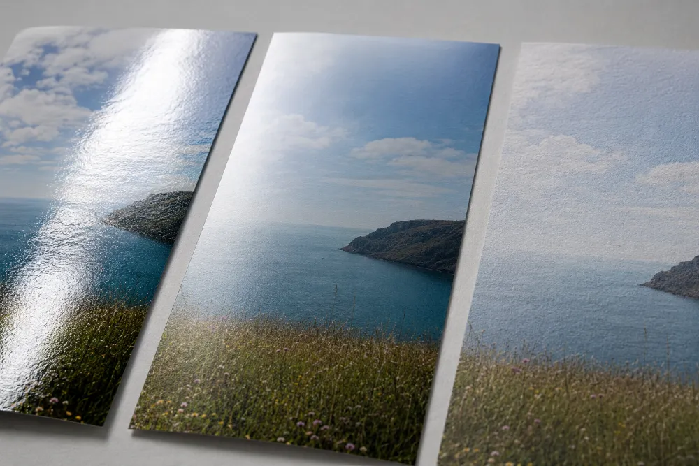 Close-up of three poster prints of the same coastal scene in gloss, satin and matte finishes, showing how gloss reflects light while matte stays glare-free