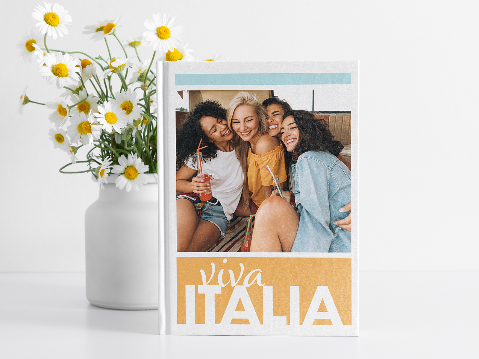 A hardcover photo book on a clean surface beside fresh daisies in a slim ceramic vase — printed on premium recycled stock